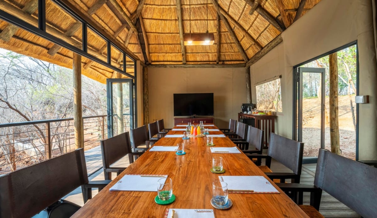 Thatched boardroom with a long timber table set for a meeting, leather chairs and glass doors onto the bush