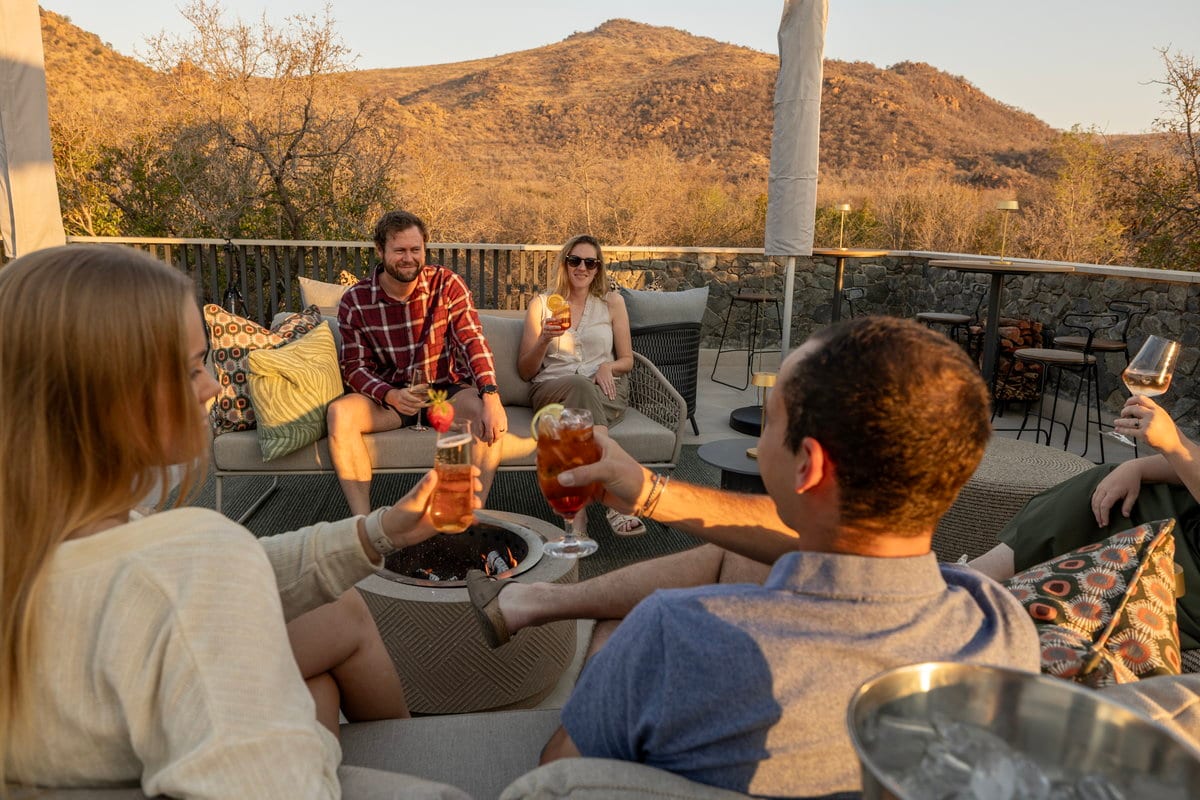 Guests sharing drinks around a fire pit on an outdoor terrace at golden hour, with hills behind