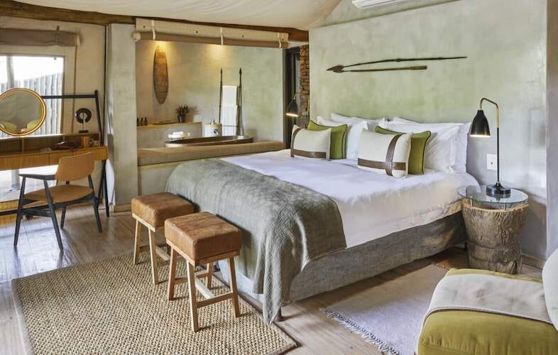 Bedroom in soft sage tones with a timber vanity, leather-topped stools and a stone bath behind the bed